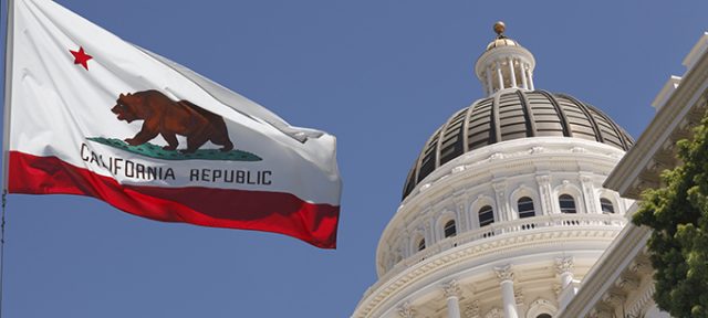 Image shows the California state flag flying in front of the capitol dome