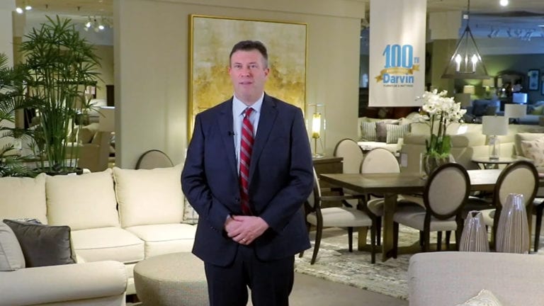 Image shows a man wearing coat and tie standing in a furniture showroom