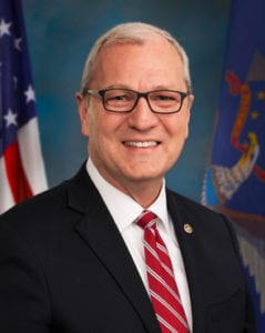 Image shows a man with white hair wearing a coat and tie