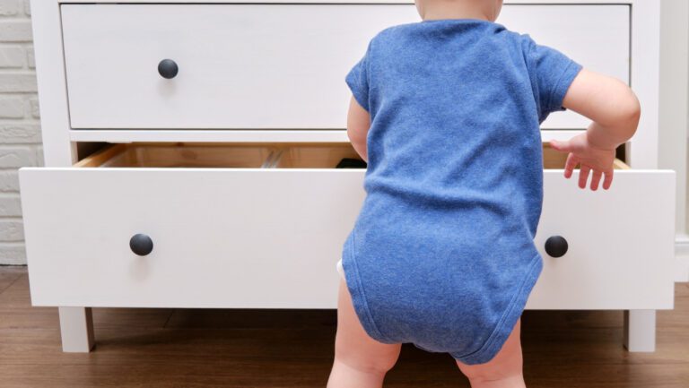 Baby standing in front of dresser with one drawer opened.