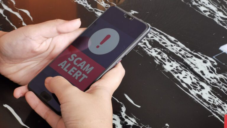 a person holding a phone with the words SCAM ALERT in red