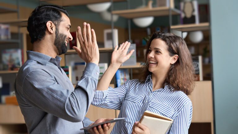 Two happy friendly diverse professionals, teacher and student giving high five standing in office celebrating success, good cooperation result, partnership teamwork and team motivation in office work.