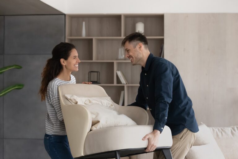 Moving day, happy buyers of fashionable furniture, bank mortgage concept. Smiling loving millennial wife and husband carry new armchair into living room, making home design improvements feel satisfied