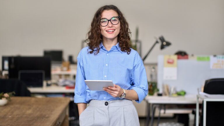 Young smart smiling professional business woman, happy confident 30s female company worker or sales manager holding digital tablet standing in modern office working, looking at camera, portrait.