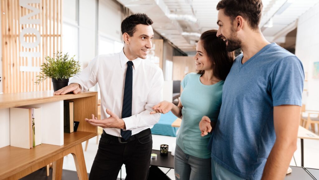 The furniture store manager demonstrates furniture to the buyers. A guy and a girl are listening with a good mood to the seller.