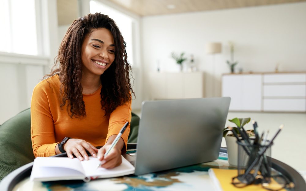 Smiling young African American woman sitting at desk working on laptop taking notes .