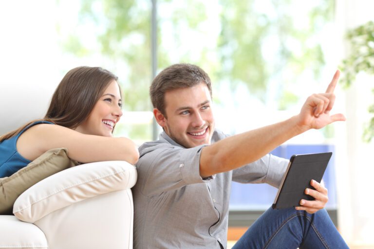Happy couple sitting and planning new decoration with a tablet on line at home with a window in the background