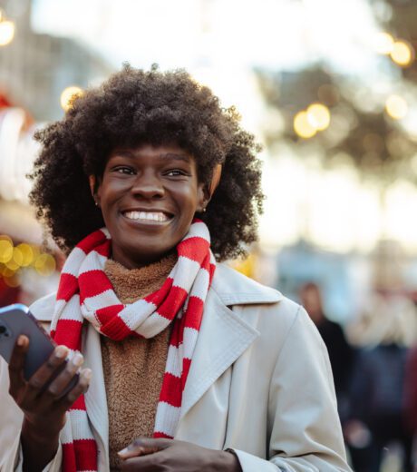 A cheerful woman with a bright scarf enjoys her smartphone at a vibrant street market during the holiday season. She smiles warmly, capturing the festive atmosphere around her.