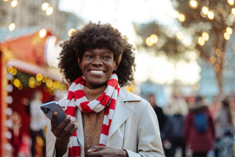 A cheerful woman with a bright scarf enjoys her smartphone at a vibrant street market during the holiday season. She smiles warmly, capturing the festive atmosphere around her.