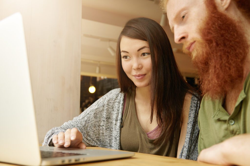 Portrait of two students using laptop while having lunch after classes at college: smiling pretty girl showing her bearded friend photographs from her trip with finger on touchpad. Film effect