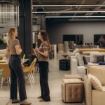 Two individuals engaged in conversation within a spacious, stylish furniture showroom, surrounded by a variety of sofas, chairs, and tables, showcasing modern interior design and decor elements.