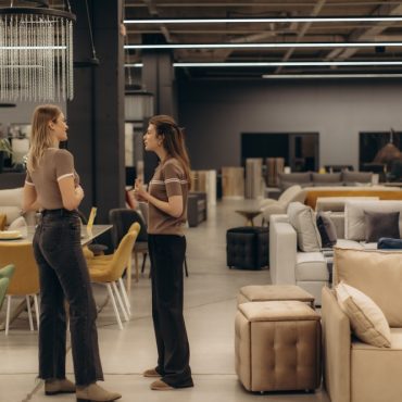 Two individuals engaged in conversation within a spacious, stylish furniture showroom, surrounded by a variety of sofas, chairs, and tables, showcasing modern interior design and decor elements.
