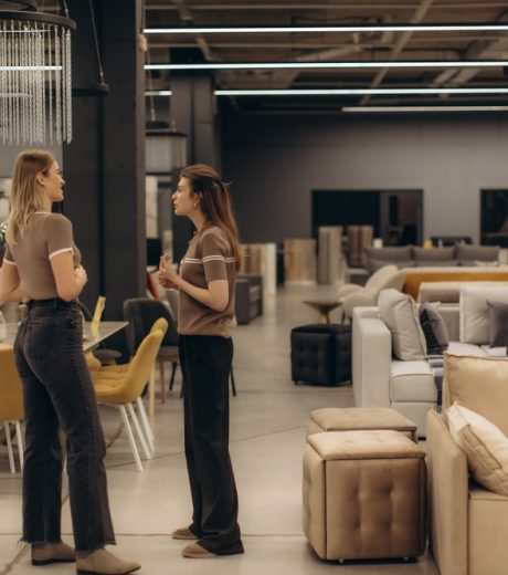 Two individuals engaged in conversation within a spacious, stylish furniture showroom, surrounded by a variety of sofas, chairs, and tables, showcasing modern interior design and decor elements.