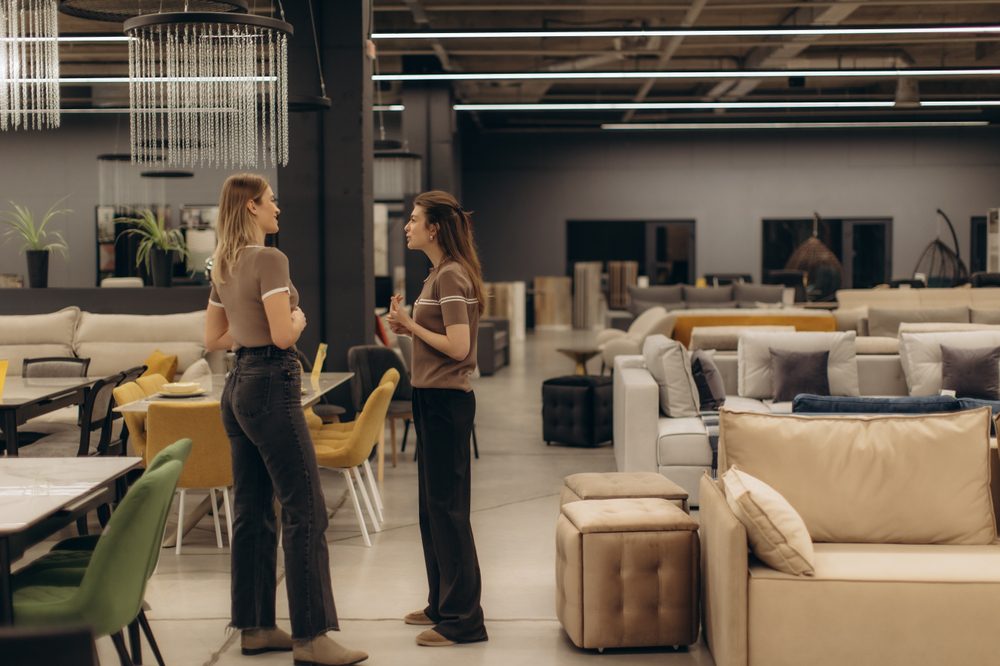 Two individuals engaged in conversation within a spacious, stylish furniture showroom, surrounded by a variety of sofas, chairs, and tables, showcasing modern interior design and decor elements.