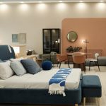 Modern furniture store interior displaying contemporary bed with pillows and blanket in foreground, stylish dining table with chairs and sofa set in background