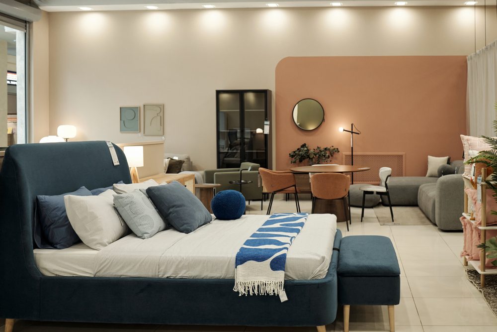Modern furniture store interior displaying contemporary bed with pillows and blanket in foreground, stylish dining table with chairs and sofa set in background