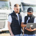 Two truck drivers discussing transportation logistics, fleet management, and shipping details using a laptop at a trucking depot