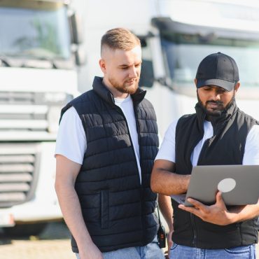 Two truck drivers discussing transportation logistics, fleet management, and shipping details using a laptop at a trucking depot