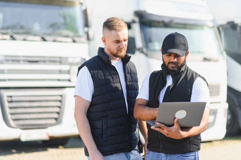 Two truck drivers discussing transportation logistics, fleet management, and shipping details using a laptop at a trucking depot