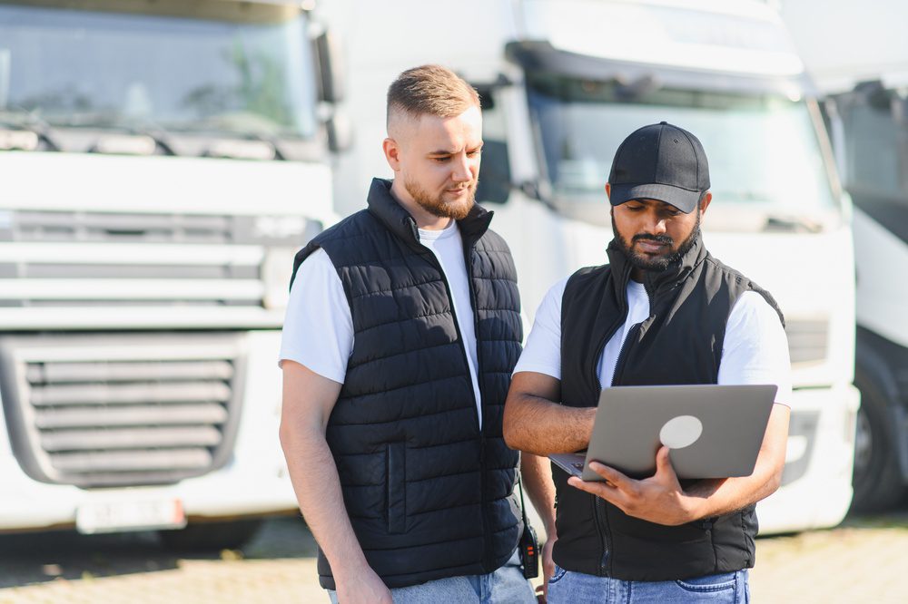 Two truck drivers discussing transportation logistics, fleet management, and shipping details using a laptop at a trucking depot